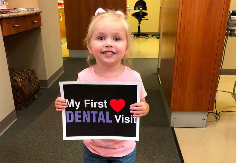 little girl holding a sign that says 'My First Dental Visit'